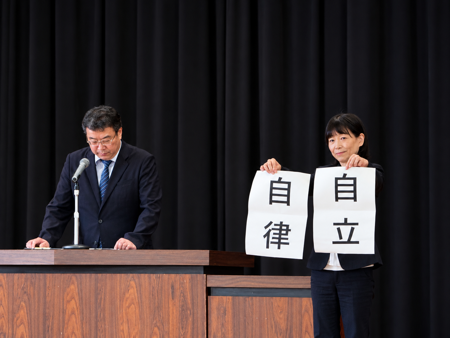 演台で話す校長先生と、「自立」「自律」と書かれた紙を掲げる教職員
